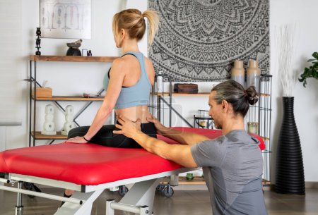 Side view of focused physiotherapist with ponytail examining lower back of female patient in activewear sitting on red stretcher during treatment in modern clinic with shelves and wall tapestry on background