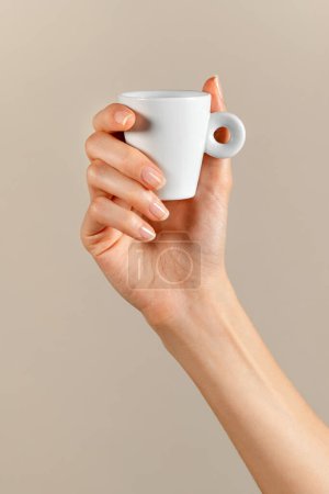 Foto de Taza femenina anónima con mini café blanco de cerámica con asa anillo en dedos delgados manicura mano contra fondo beige en luz - Imagen libre de derechos
