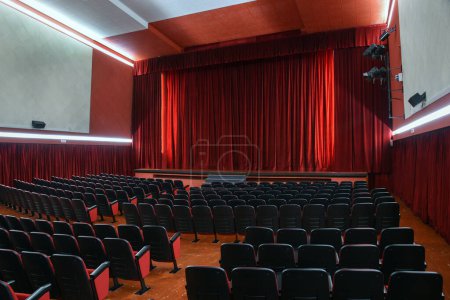 Foto de Interior de teatro vacío con filas de cómodos sillones bajo luces iluminadoras, mientras que las cortinas de color rojo brillante que cuelgan a lo largo de las paredes y desde el techo del escenario - Imagen libre de derechos