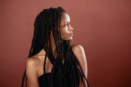 Foto de Retrato de una joven afroamericana reflexiva con largas trenzas afro y hombros desnudos mirando hacia otro lado mientras está sentada en la habitación sobre un fondo marrón en la luz - Imagen libre de derechos