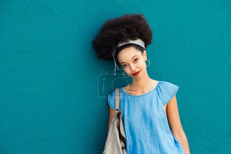 Foto de Retrato de joven marroquí sonriente maquillada con diadema de pelo afro mirando a la cámara mientras está de pie con el bolso contra el fondo azul a la luz del día - Imagen libre de derechos