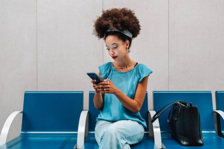 Foto de Mujer marroquí joven concentrada con cabello afro y diadema mirando la pantalla del teléfono móvil mientras está sentada con el bolso en la estación de autobuses bajo las luces y buscando mensajes de texto - Imagen libre de derechos