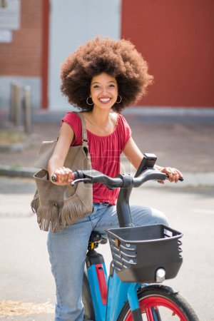 Foto de Mujer afroamericana positiva en ropa casual con peinado afro y bolso sonriendo y mirando a la cámara mientras monta bicicleta eléctrica en la calle de la ciudad - Imagen libre de derechos