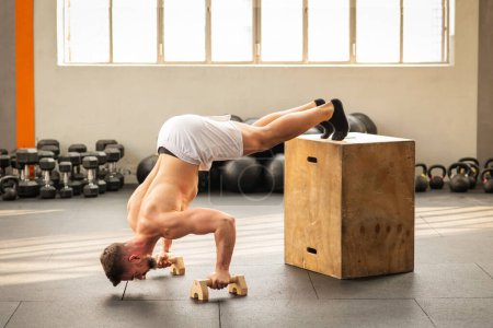 Foto de Vista lateral del fuerte musculoso macho que realiza ejercicio de empuje hacia arriba en cubo de madera en el gimnasio moderno - Imagen libre de derechos