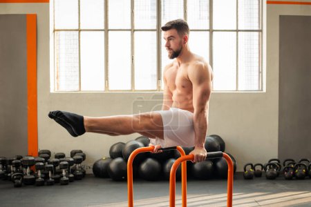 Foto de Vista lateral de los músculos masculinos haciendo L sentarse en barras paralelas ejercicio de calistenia en el gimnasio moderno - Imagen libre de derechos