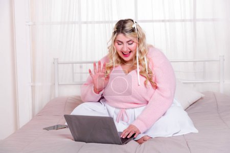 Foto de Señora con curvas positivas en suéter sentado en la cama cómoda saludando la mano mientras se utiliza el ordenador portátil para hacer videollamada en una habitación acogedora - Imagen libre de derechos