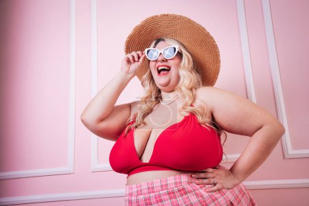 Foto de Emocionada modelo femenina con sonrisa dentada con sombrero y gafas de sol de pie detrás de fondo rosa en el estudio - Imagen libre de derechos