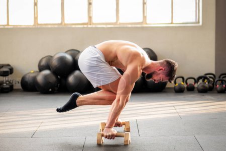 Foto de Vista lateral del macho musculoso fuerte haciendo ejercicio de calistenia de planche Tuck en el suelo en el gimnasio con paralelos de madera - Imagen libre de derechos