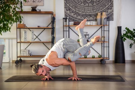 Cuerpo completo de macho enfocado en ropa deportiva practicando Kakasana brazo equilibrio yoga pose sobre estera en habitación iluminada durante el día