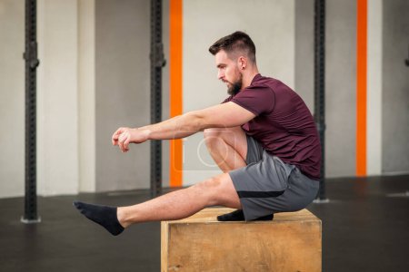 Side view of strong bearded adult male athlete in sportswear looking away while sitting on box doing calisthenics pistol squat exercise in daylight against modern building wall