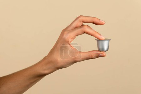 Close-up of a human hand elegantly presenting a coffee capsule, isolated on a beige backdrop