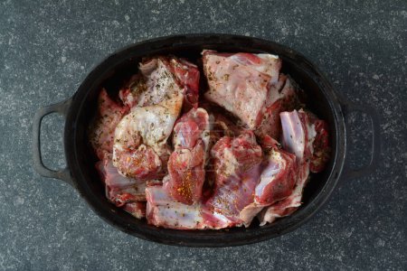 Seasoned lamb meat in black iron pot, ready to roast, over dark stone background, top view