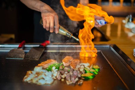 manos del chef con espátula sobre teppanyaki. cocinar verduras carne y mariscos en la mesa de parrilla de hibachi caliente. Cocina tradicional japonesa. Espectáculo de Teppan con flambe