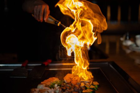 Photo for Chef's hands with spatula over teppanyaki. cooking vegetables meat and seafood on hot hibachi grill table. Traditional Japanese Cuisine. Teppan show with flambe - Royalty Free Image