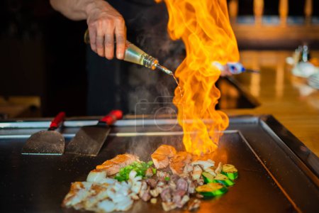 Photo for Chef's hands with spatula over teppanyaki. cooking vegetables meat and seafood on hot hibachi grill table. Traditional Japanese Cuisine. Teppan show with flambe - Royalty Free Image