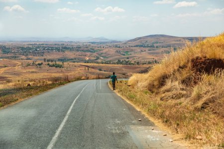 Foto de Antsirabe, Madagascar 19 de octubre de 2023. Carretera de Morondava a Antsirabe a través de pastizales vacíos y pueblos en el centro de Madagascar. hombre solitario en la distancia camina a lo largo del lado de la carretera - Imagen libre de derechos