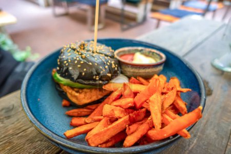 Schwarzer Burger mit Pommes frites auf hölzernem Hintergrund serviert auf einem blauen Keramikteller