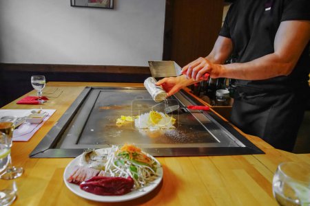 chef's hands with spatula over teppanyaki. cooking vegetables meat and seafood on hot hibachi grill table. Traditional Japanese Cuisine. Teppan show