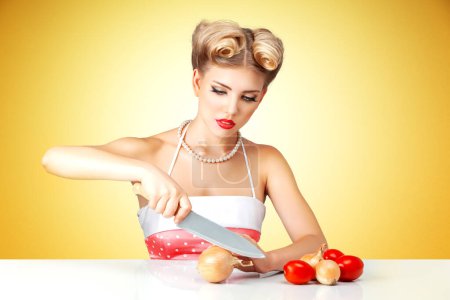 Photo for Young blonde housewife chopping onion on table in kitchen. Retro classic 50s style photoshoot. - Royalty Free Image