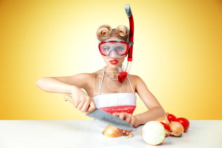 Téléchargez les photos : Young blonde housewife chopping onion in diving mask to protect her eyes. Retro classic 50s style photoshoot. - en image libre de droit