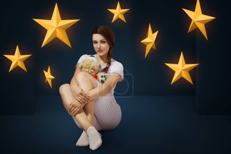 Photo for Young brunette woman wearing pajama hugging teddy bear in the bedroom decorated with stars on background - Royalty Free Image