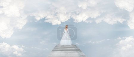 Photo for Young lady woman in white romantic wedding dress walking along fantasy bridge in cloudy skies - Royalty Free Image