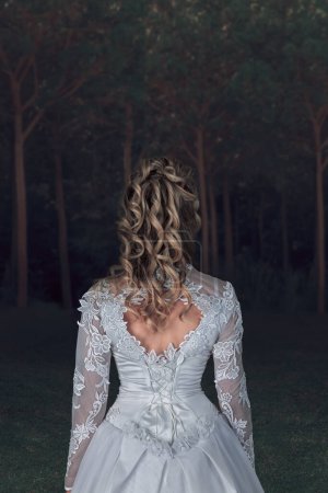 Photo for Back view of a woman in white wedding dress standing in moody dark forest. Rear view - Royalty Free Image