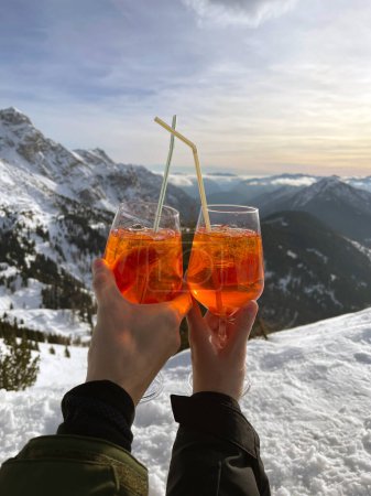 Foto de Una pareja sostiene vasos de Aperol en copas de vidrio con pajitas en una cima de la montaña. Vistas panorámicas de las montañas y la nieve. Manos de amantes en la montaña. - Imagen libre de derechos