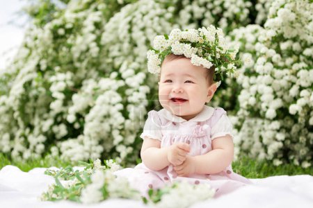 Foto de Dulce niña sonriente con corona de flores de espirea en vestido rosa, sobre hierba verde cerca de arbustos de espirea en el primer día soleado de verano - Imagen libre de derechos