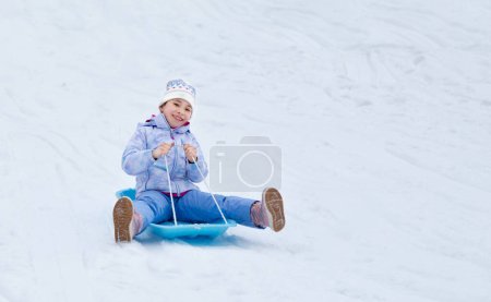 Concept of childhood, sledding in winter. Happy little girl is rolling down the hill on a sled. Happy holidays.