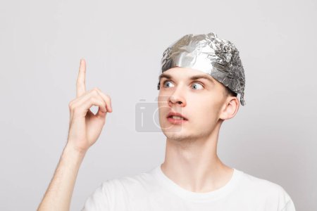 Photo for Portrait of crazy young man wearing tin foil hat pointing his finger up. Conspiracy theories and paranoya concept. Studio shot on gray background - Royalty Free Image
