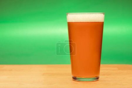 Foto de Full shaker pint glass of amber ale or beer on wooden table with green background - Imagen libre de derechos