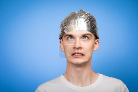 Photo for Portrait of anxious young man wearing tin foil hat looking up in panic. Conspiracy theories and paranoya concept. Studio shot on blue background - Royalty Free Image