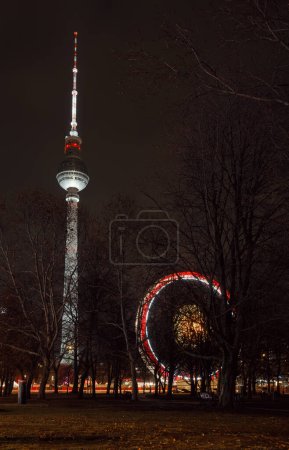 Photo for Berlin, Germany - December 12, 2018: Spinning illuminated Ferris Wheel and Fernsehturm Television Tower seen thru trees in Central Berlin - Royalty Free Image