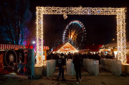 Photo for Berlin, Germany - December 12, 2018: Visitors entering the Christkindlmarkt (Christmas market) thru illuminated gate. Illuminated Gluhwein (hot mulled wine) stall and Ferris Wheel are seen in background - Royalty Free Image