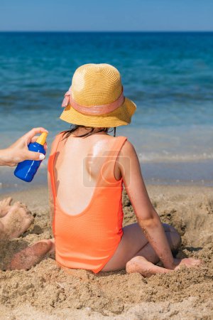Photo for Mother spraying sunscreen on her daughter's back while she playing on a beach - Royalty Free Image
