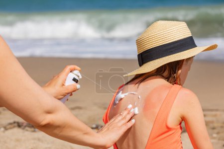 Photo for Mother spraying panthenol foam on her daughter's sunburnt back on a beach - Royalty Free Image