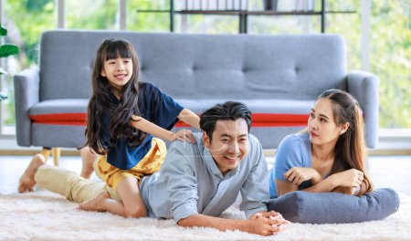 Photo for Millennial Asian lovely happy family father mother laying down on carpet floor while young daughter girl sitting piggy back on dad smiling laughing playing together in living room at home in weekend. - Royalty Free Image