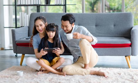 Foto de Millennial asiática feliz familia padre bebiendo café caliente, mientras que la madre y dauthger sentado en el acogedor piso de la alfombra sonriendo ayudar a jugar streaming juego remoto en línea en la televisión en casa sala de estar. - Imagen libre de derechos
