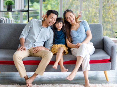 Photo for Millennial Asian happy family father mother and little young child girl sitting smiling looking at camera together on cozy sofa couch showing two fingers peace sign taking photo in living room home. - Royalty Free Image