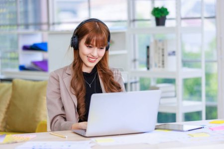 Photo for Asian professional successful young female businesswoman creative graphic designer in casual outfit sitting smiling wearing headphone listening to music playlist while working typing laptop computer. - Royalty Free Image