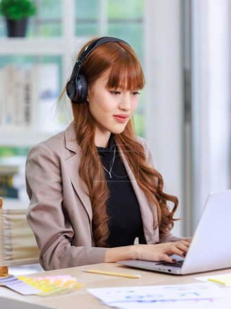 Photo for Asian professional successful young female businesswoman creative graphic designer in casual outfit sitting smiling wearing headphone listening to music playlist while working typing laptop computer. - Royalty Free Image