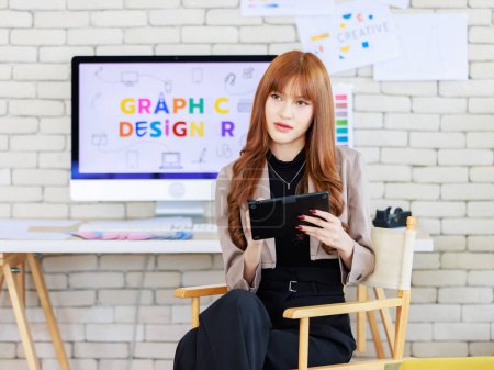 Photo for Asian professional successful young female creative graphic designer in casual fashionable blazer suit sitting smiling holding tablet computer working with computer in workspace in company office - Royalty Free Image
