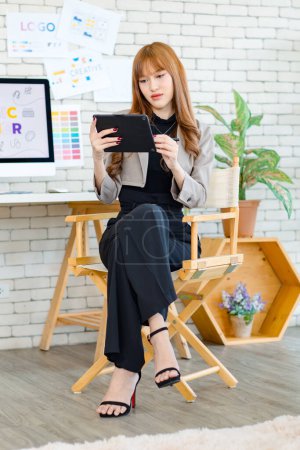 Photo for Asian professional successful young female creative graphic designer in casual fashionable blazer suit sitting smiling holding tablet computer working with computer in workspace in company office - Royalty Free Image