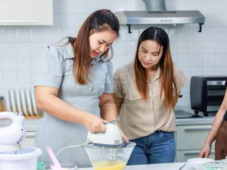 Asian happy professional female pastry bakery baker chef housewife with close friends standing smiling helping using eggs whisk beater machine mixing flour preparing dough in full decorated kitchen.