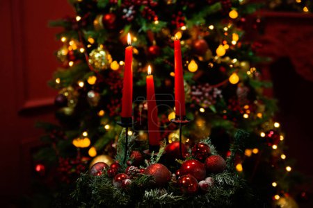 Téléchargez les photos : Trois bougies rouges allumées dans l'obscurité contre le bokeh d'un sapin de Noël, lumières et décorations. Concept de Noël et nouvelle année. - en image libre de droit