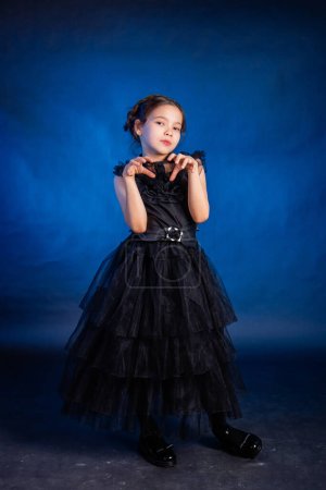 A little girl in a black dress with a pigtail hairstyle on her head poses, isolated on a dark background with blue backlight.