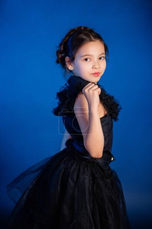 Portrait of a little girl in a black dress with a pigtail hairstyle on her head poses, isolated on a dark background with blue backlight. 