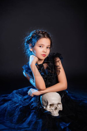 Foto de Una niña con un vestido negro con un peinado de coleta en la cabeza posa sentada con un cráneo en las manos, aislada sobre un fondo oscuro con luz de fondo azul. - Imagen libre de derechos