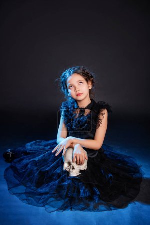 Ein kleines Mädchen in einem schwarzen Kleid mit einer Zopf-Frisur auf dem Kopf posiert sitzend mit einem Totenkopf in den Händen, isoliert auf einem dunklen Hintergrund mit blauer Hintergrundbeleuchtung.